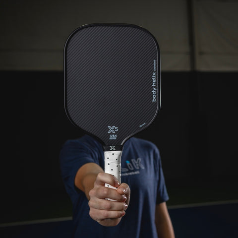 Person holding a black pickleball paddle with 'body helix' branding against a dark background
