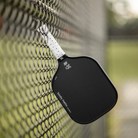 body helix X5 Black kevlar pickleball paddle with white handle hangin on  a chain-link fence